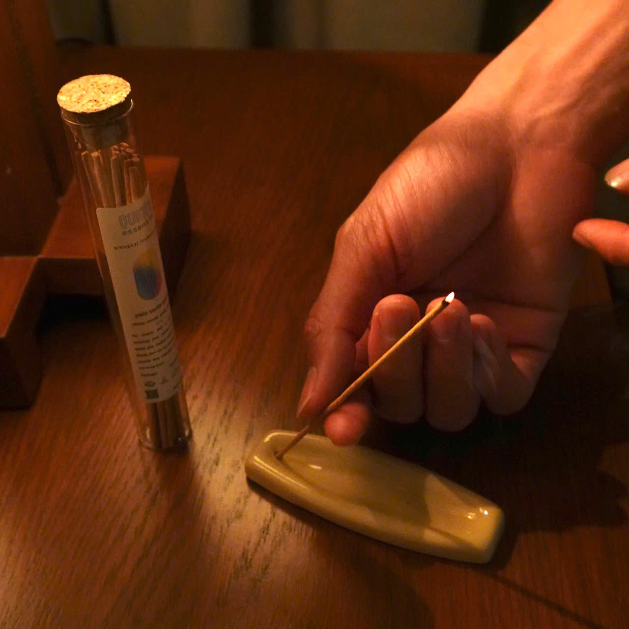 Person lighting a Palo Santo incense stick on a wooden table with a tube of Palo Santo incense sticks in the background.